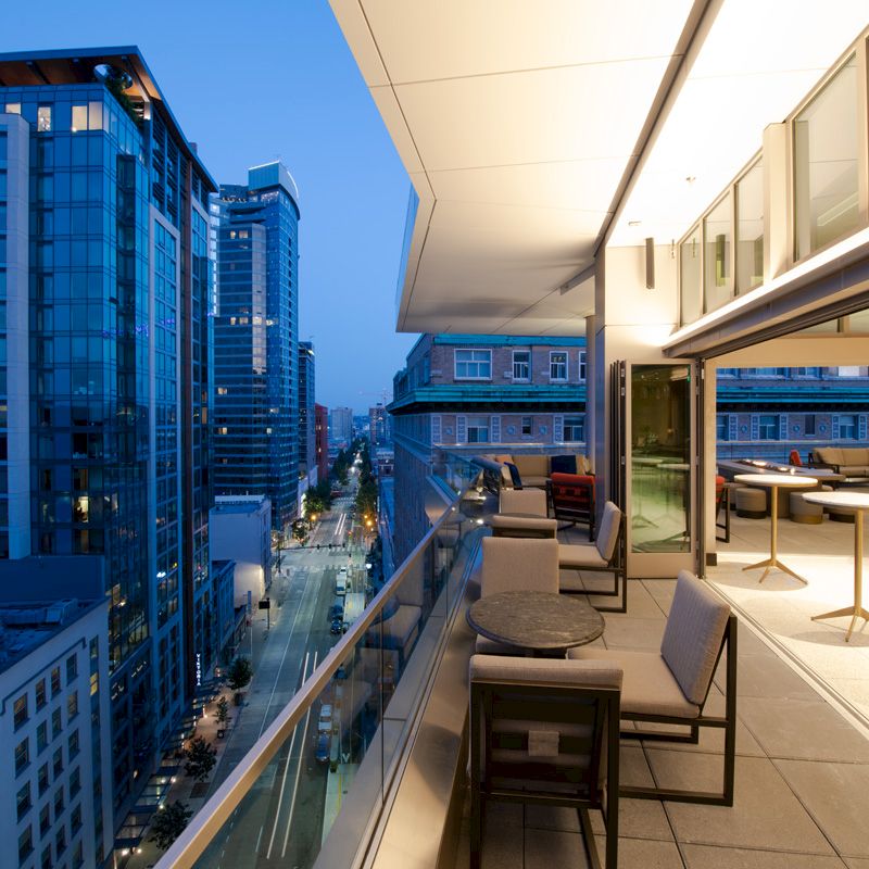 A balcony view in a high-rise building, overlooking a city street with modern glass buildings. The balcony has tables and chairs for seating.