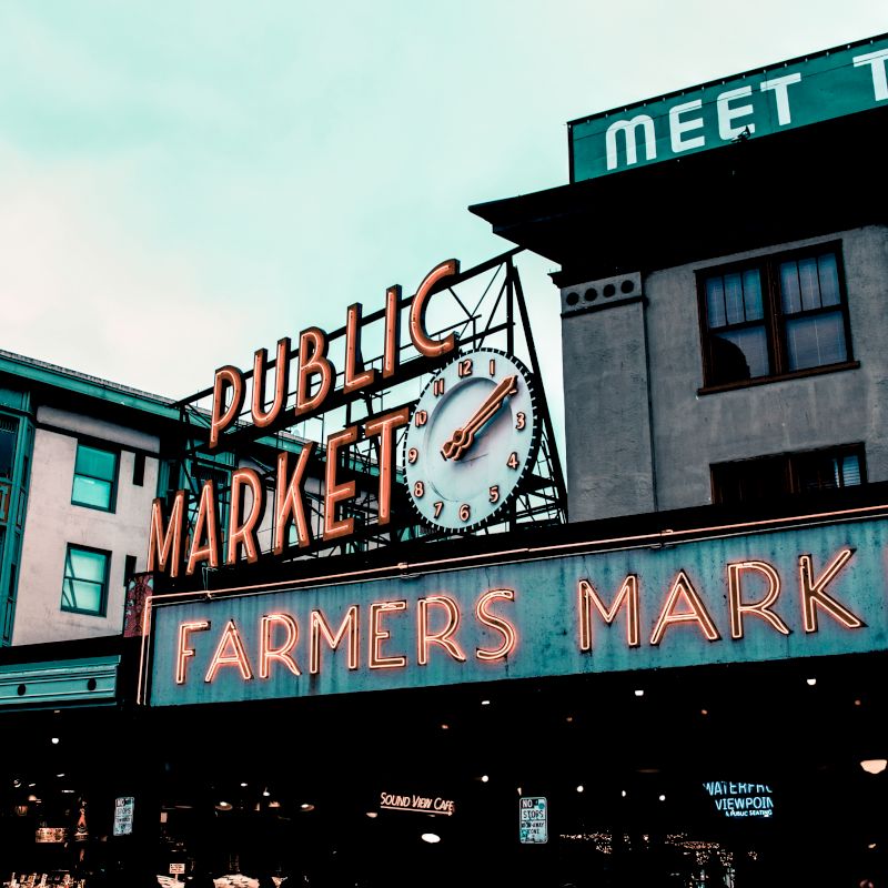 The image shows a neon sign for "Public Market" and "Farmers Market" with a clock in between, set against an urban backdrop.