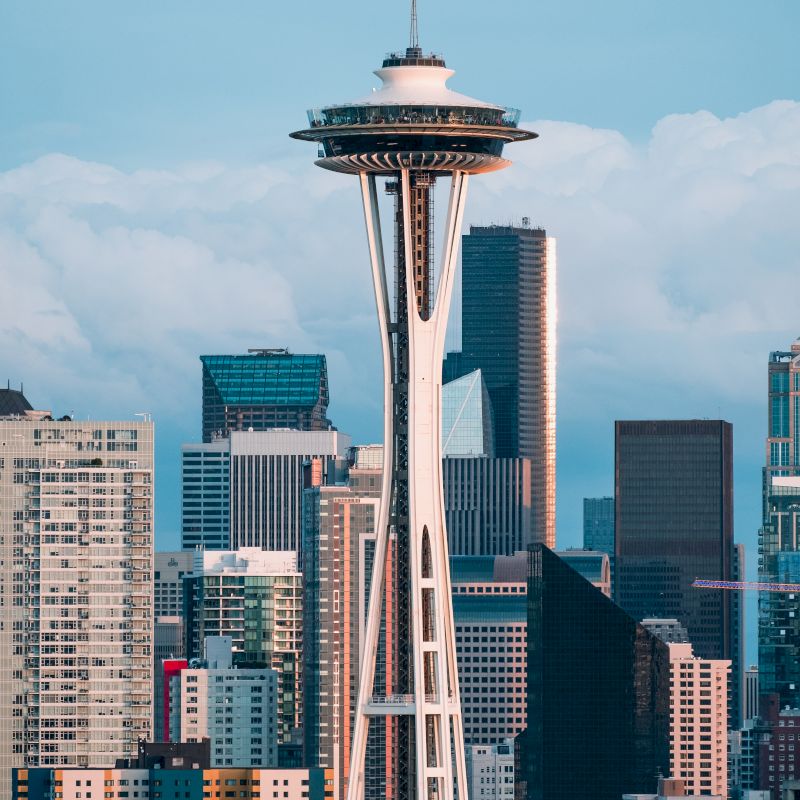 The image shows the Space Needle, a famous observation tower, with various modern and high-rise buildings in the background in a cityscape.