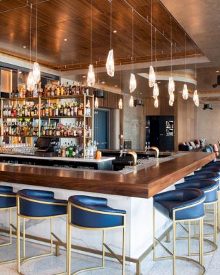 The image shows a modern bar with a variety of bottles on shelves behind the counter, surrounded by blue barstools and contemporary lighting.