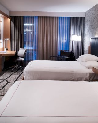 The image shows a modern hotel room with two beds, a wall-mounted TV, a desk with a chair, and stylish decor including a patterned wall and floor.