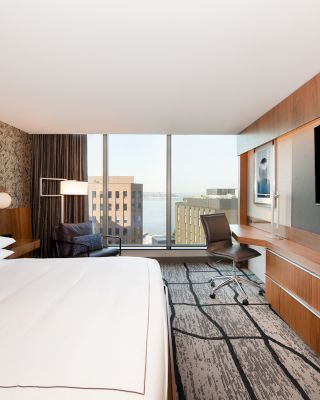 A modern hotel room features a large bed, a wall-mounted TV, a desk with a chair, a city view through floor-to-ceiling windows, and stylish decor.