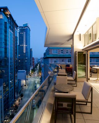 A modern balcony with seating extends from an upscale interior, overlooking a cityscape with tall buildings and a busy street below at dusk.