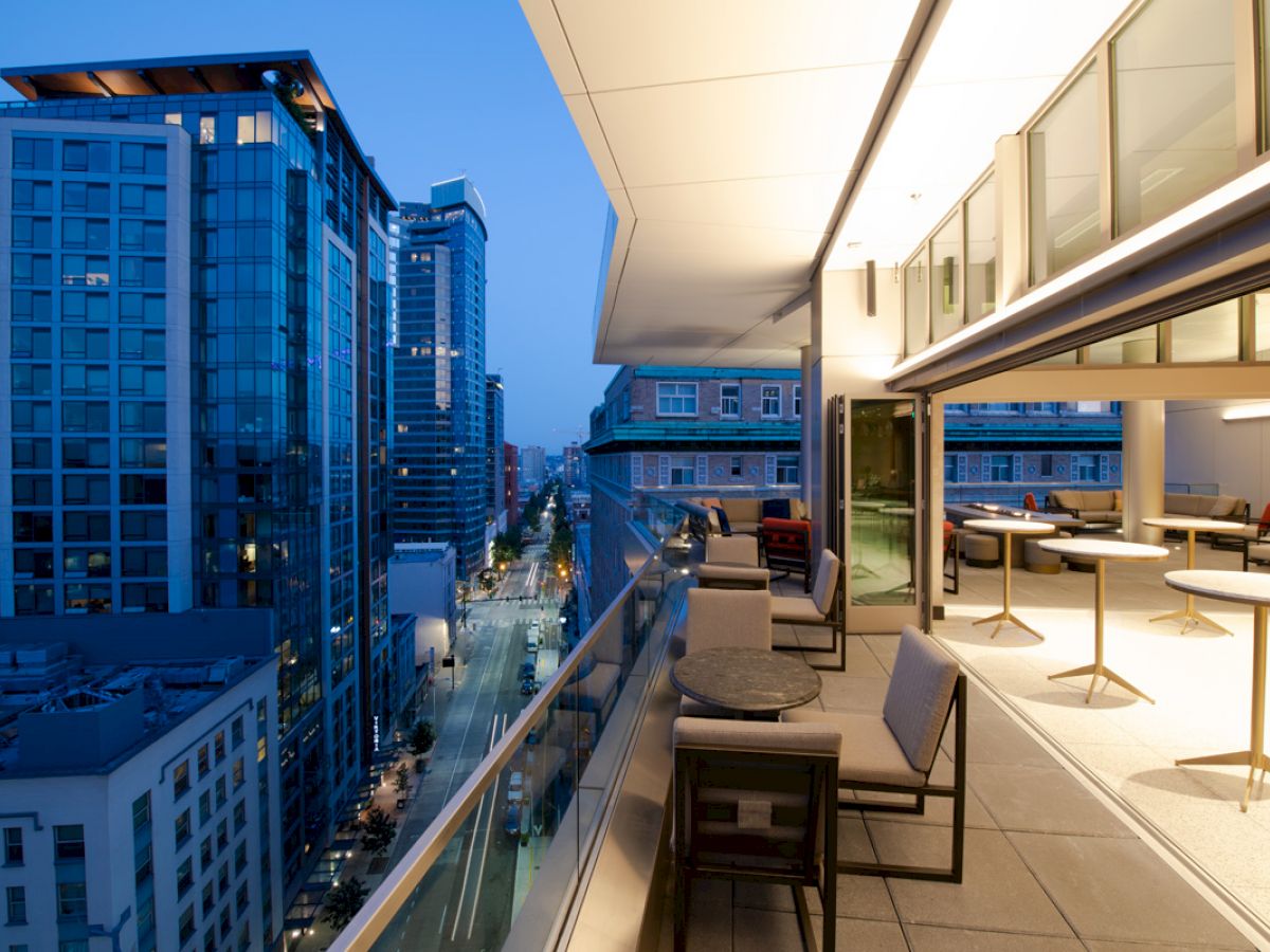 The image shows a modern balcony with seating, overlooking a cityscape with tall buildings at dusk. The interior space is also visible.