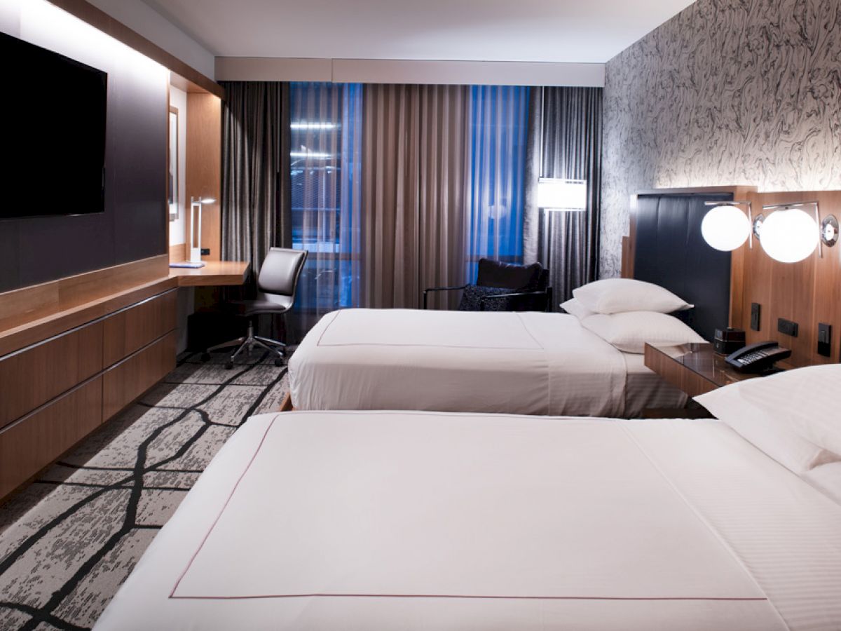 A hotel room featuring two double beds, a flat-screen TV, a work desk with a chair, modern lighting, and large windows with curtains.
