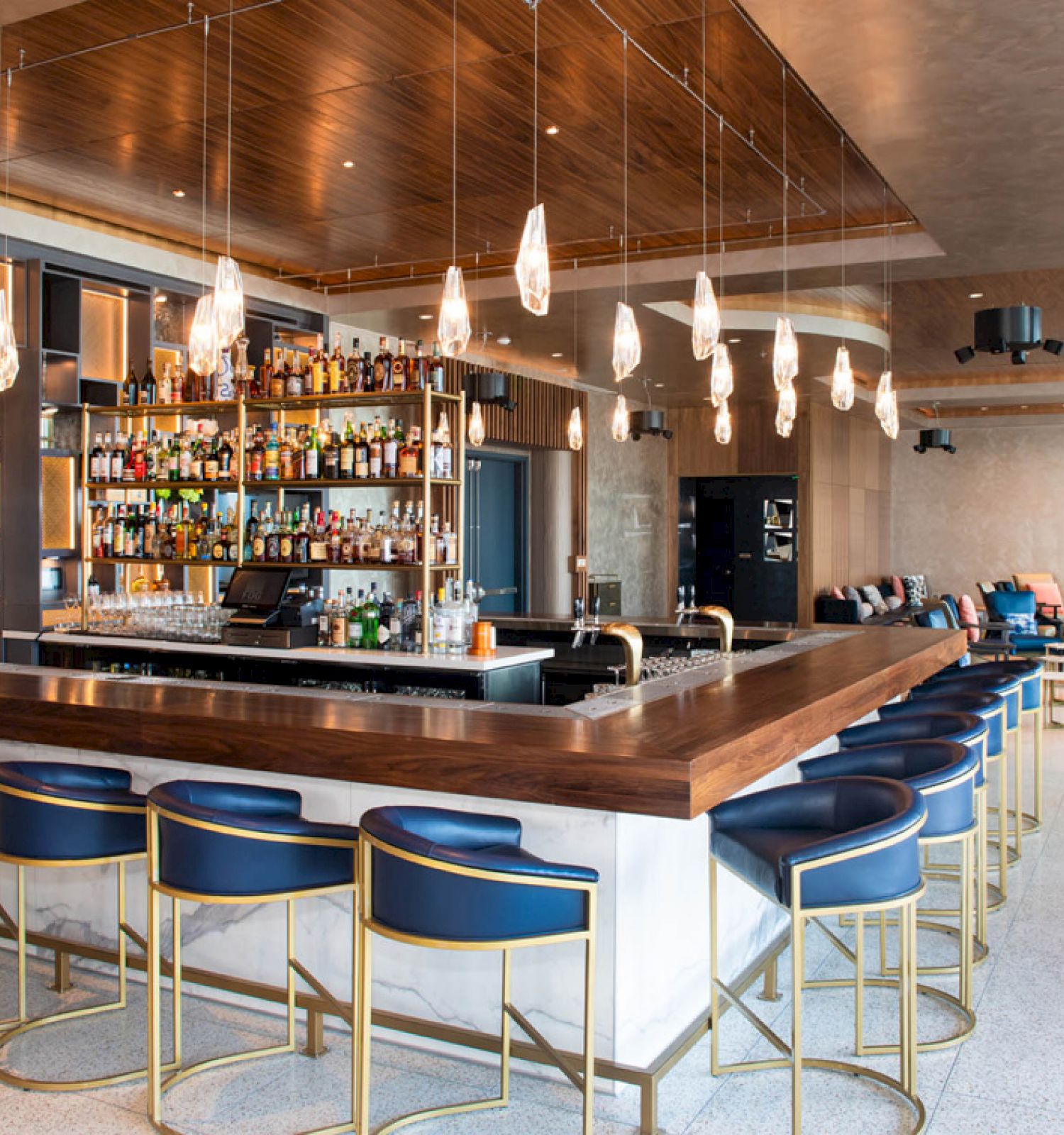 A modern bar with a well-stocked liquor display, blue barstools, and pendant lighting. The seating area has couches and tables.