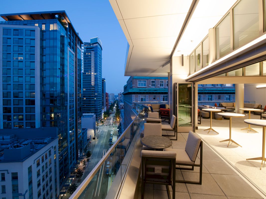 This image shows a modern cityscape viewed from a balcony with seating areas at dusk, featuring tall buildings and lighted streets below.