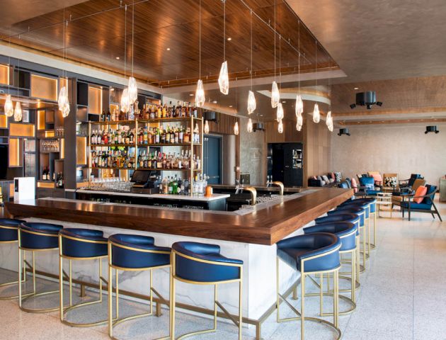 A modern bar with a large wooden counter, blue bar stools, hanging lights, a fully stocked shelf, and a seating area with tables and sofas.