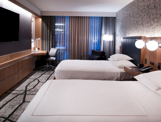 A modern hotel room with two beds, a flat-screen TV, a desk with a chair, wall-mounted lamps, and a large window with curtains.