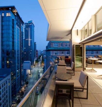 A modern cityscape view from a high-rise balcony at dusk, featuring outdoor seating and tables, with buildings and streets illuminated below.
