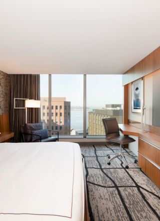 A modern hotel room with a large bed, desk, chair, big TV, and a window view of buildings and water. The decor is contemporary with wood accents.