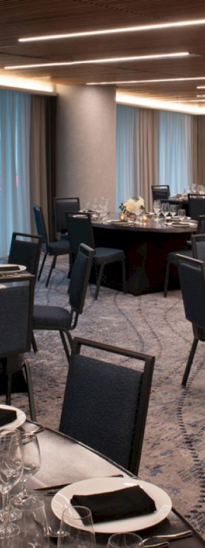 A spacious, elegant event room is set up for a formal dinner with round tables, chairs, place settings, and floral centerpieces under modern lighting.