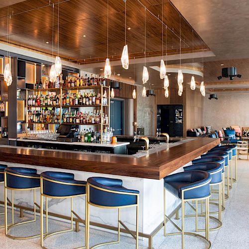 The image shows a modern bar with high chairs, a well-stocked liquor shelf, and stylish hanging lights. The space is elegant and open-ended.