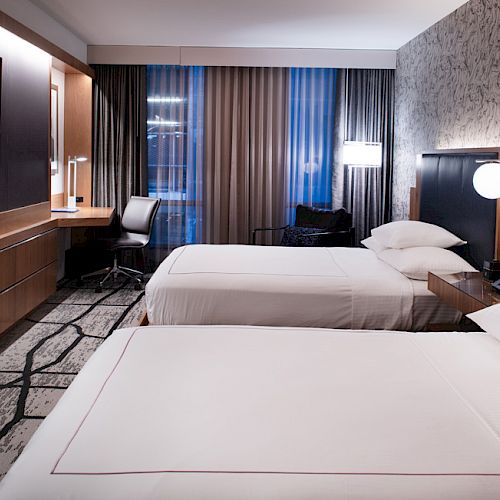 A modern hotel room with two beds, a flat-screen TV, a desk, chairs, and floor-to-ceiling windows with curtains.