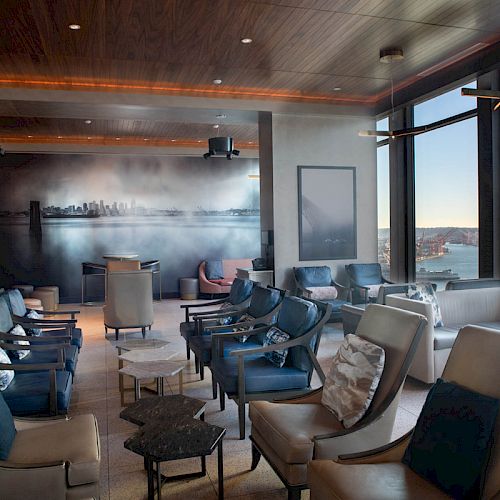 A modern lounge area with comfortable seating, large windows offering a view of a body of water, and a wall mural of a city skyline.