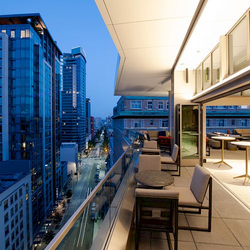A modern cityscape during twilight featuring high-rise buildings, a spacious balcony with seating, and large windows opening to a well-lit interior.