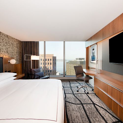 A modern hotel room with a large bed, wall-mounted TV, desk, and a window with a city view featuring tall buildings.