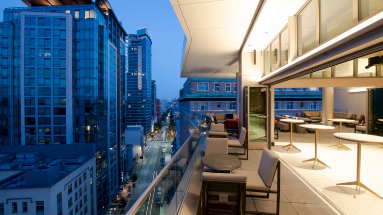 A modern, high-rise balcony overlooking a city street with buildings on both sides at dusk, featuring outdoor seating and tables.