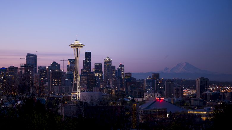The image depicts Seattle's skyline at dusk, featuring the Space Needle and Mount Rainier in the background, under a clear sky.