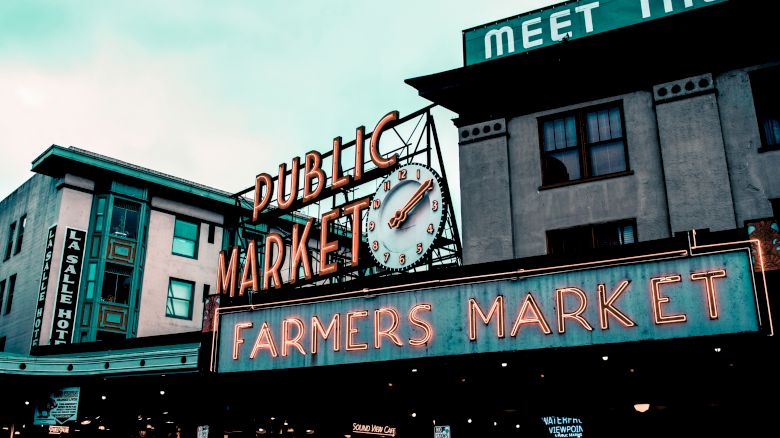 The image shows a neon sign for "Public Market" and "Farmers Market" with a clock, and nearby buildings with signs, likely a famous market location.