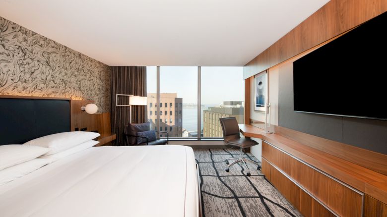 A modern hotel room with a large bed, a desk with a chair, a flat-screen TV, and a window with a cityscape view.