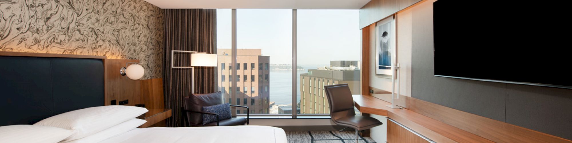 A modern hotel room with a large bed, a desk and chair, a wall-mounted TV, and a city view through large windows ends the sentence.