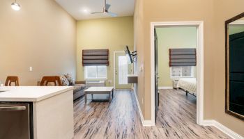 The image shows a modern, open living space with a kitchen area, living room, and a connected bedroom with a bed and nightstand visible through an open door.