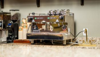 The image shows a coffee station with an espresso machine, a coffee grinder, disposable cups, and a faucet on a marble countertop.