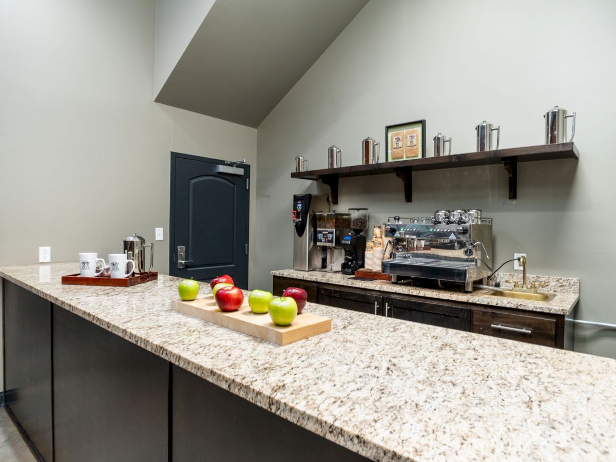 A modern kitchen setup with a granite countertop, coffee machine, apples on a tray, and kitchenware on the shelves.