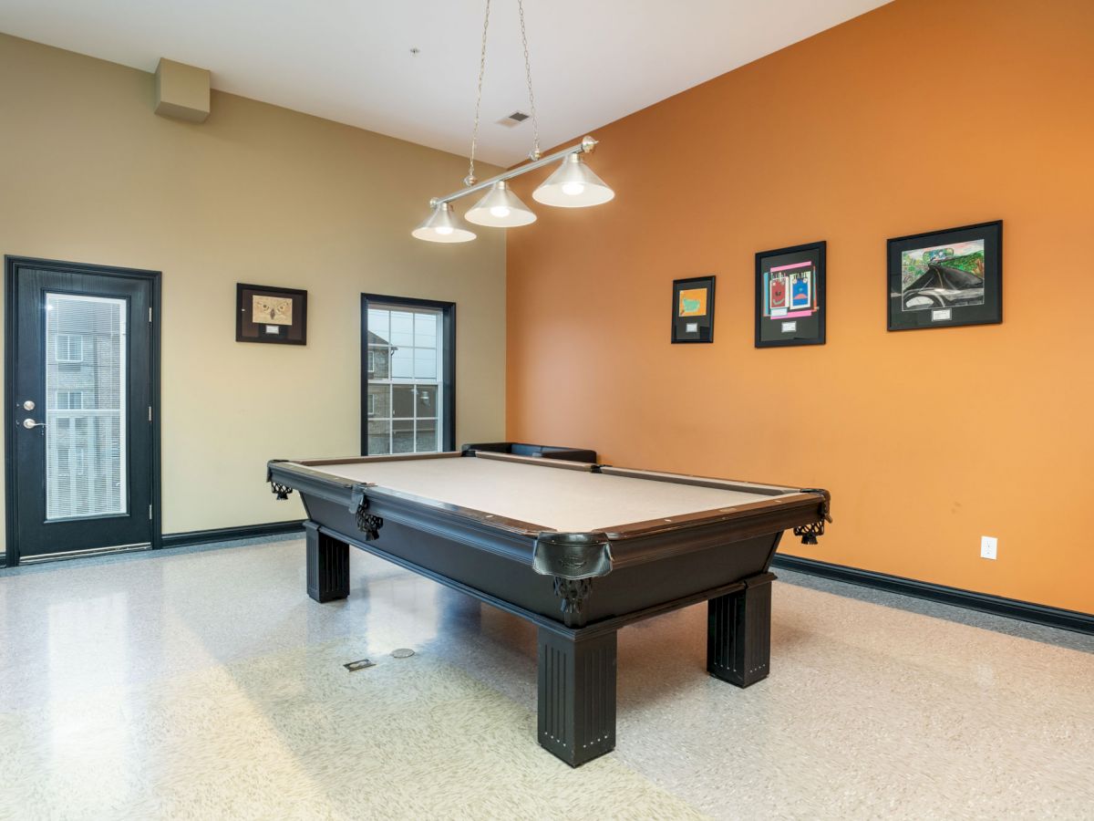 A room with a billiards table, framed pictures on an orange wall, and a door and window on the adjacent wall. Three hanging lights above the table.